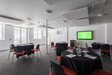 Four cabaret style tables are in the Courtyard room with a digital screen at the front of the room. The walls are white and there's a white beam in the centre of the room.