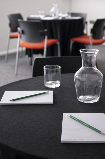 A photograph of Bishopsgate Institute's Courtyard Room in central London. The image is focused on a table top with pens and paper on it and a glass of water.