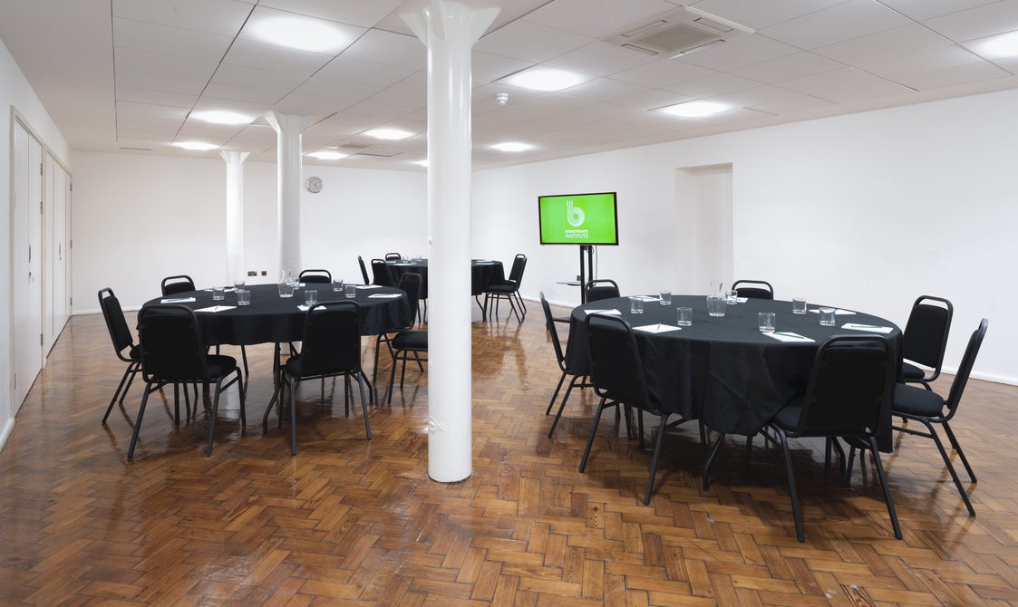 A photograph of Bishopsgate Institute's Goss Room, with the tables set up in a cabaret style with chairs around them. There is a digital screen at the back of the room.