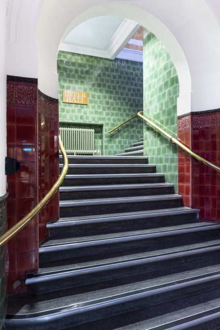 A staircase at Bishopsgate Institute. The stairs are black with gold bannisters. There are red tiles on the wall leading up to another staircase with green tiles.