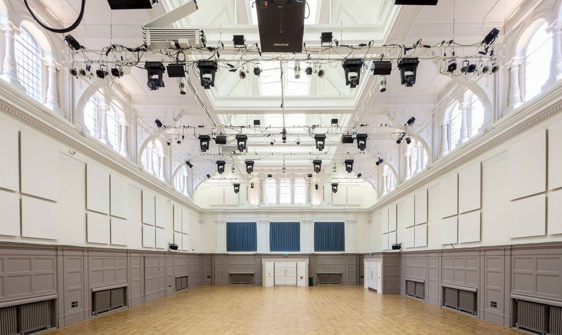 A photograph of Bishopsgate Institute's Great Hall in central London. The walls are white and grey with a wooden floor. There is a lighting rig pointed towards a stage.