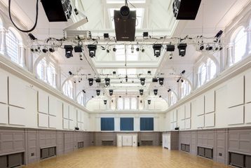 A photograph of Bishopsgate Institute's Great Hall in central London. The walls are white and grey with a wooden floor. There is a lighting rig pointed towards a stage.