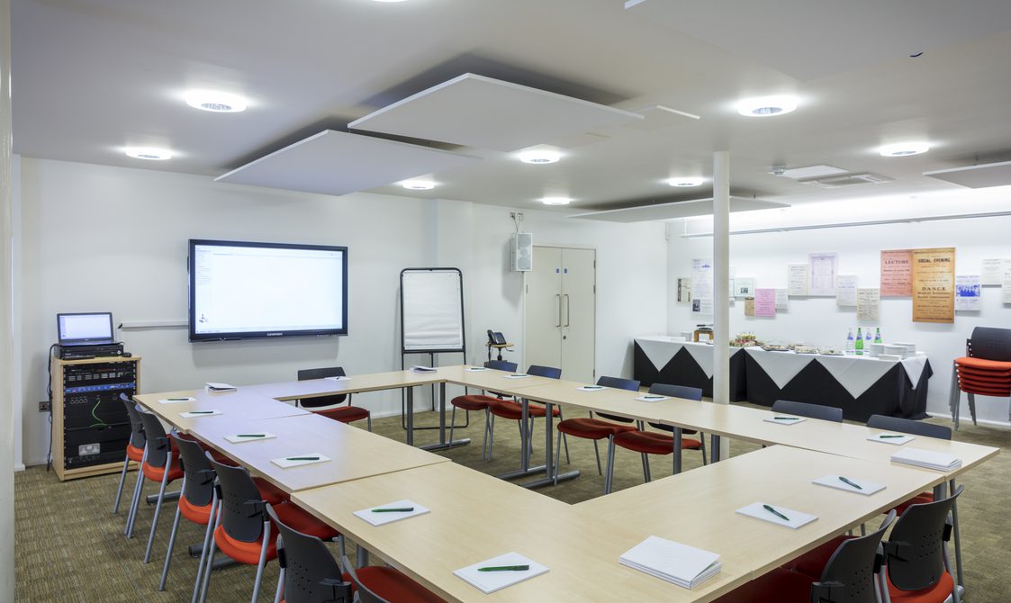 A photograph of the Courtyard Room, which has the tables set up in a roundtable style, with chairs around it. There is a screen at the front of the room, and a catering table to the side.