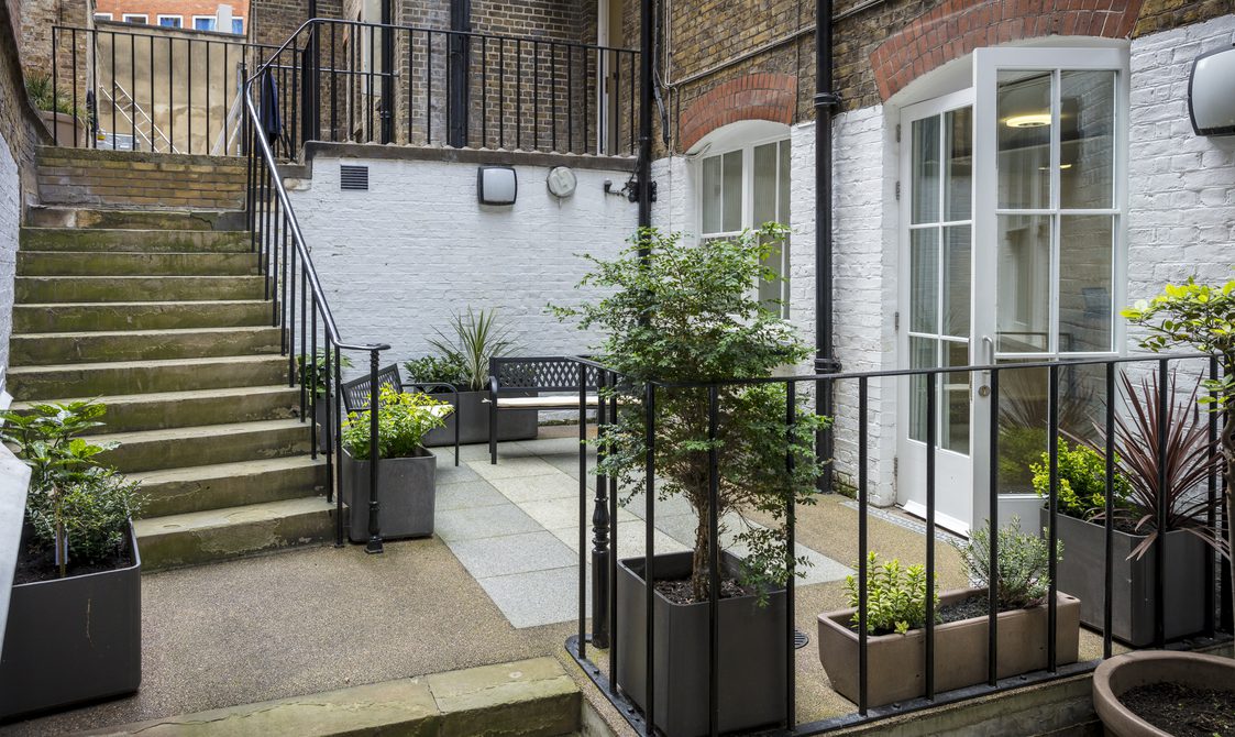 A photograph of the outside of the Courtyard Room. There is a small patio with plants on it, and stairs leading up to the main road.