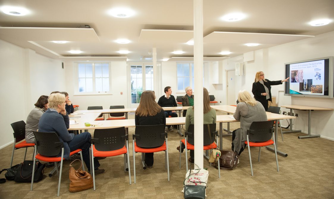 A photograph of the Courtyard Room, which has people sitting round a rectangular table and someone at the front presenting, with a screen behind them.