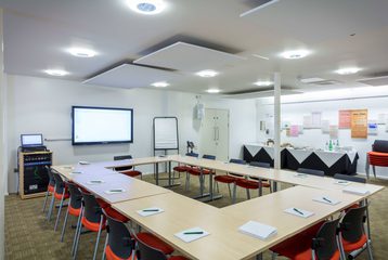 A photograph of Bishopsgate Institute's Courtyard Room. The tables are set up in a board room style with chairs around it. There is a screen on the wall and a catering table to the side of the room.
