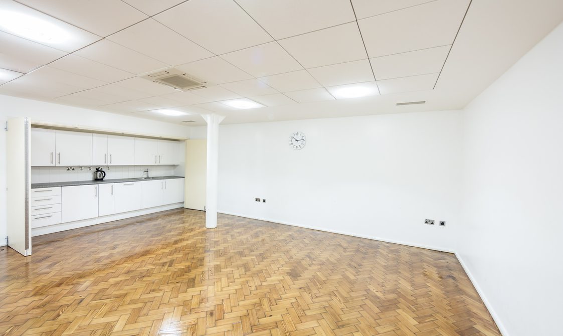 A photograph of the Goss Room. The walls are white and the floor is wooden laminate. There is a kitchenette in the room, which has a kettle on top of the work surface.