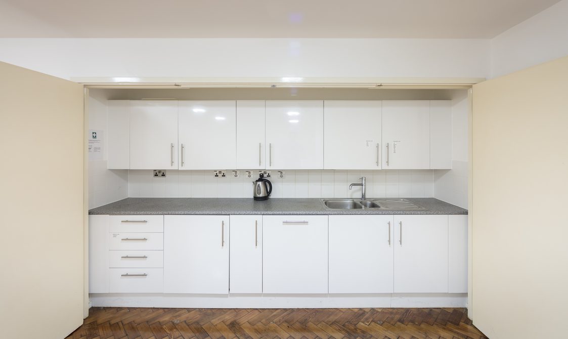 A photograph of the Goss Room. The image focuses on the kitchenette, which is white with a grey marble surface. There is a sink and kettle on the worktop.
