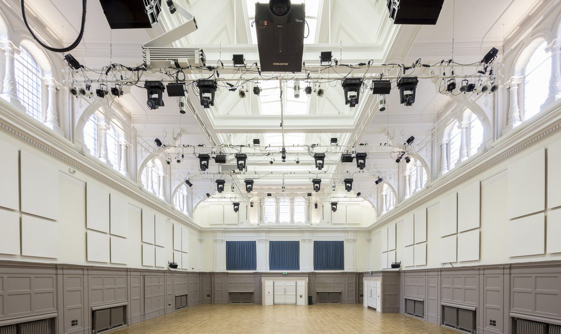 A photograph of the Great Hall with natural lights coming through the windows. There is a lighting rig, and doors at the end of the photo. The walls are painted grey and white.