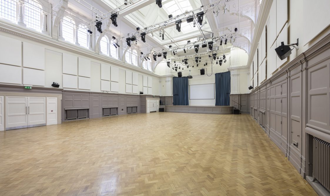 A photograph of the Great Hall with natural light coming through the windows. There is a stage with blue curtains and a lighting rig pointed at the stage.