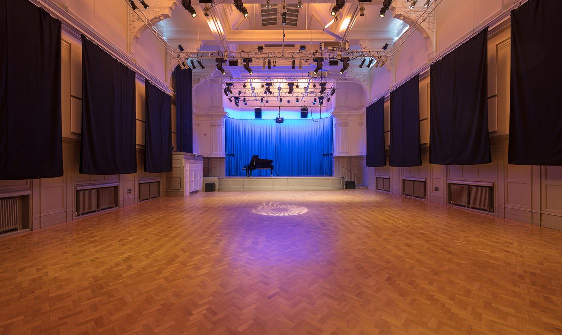A photograph of the Great Hall. There is a piano on stage with a blue curtain. There is a lighting rig with soft lighting over the stage.