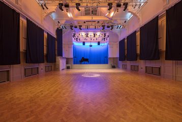 A photograph of the Great Hall. There is a piano on stage with a blue curtain. There is a lighting rig with soft lighting over the stage.