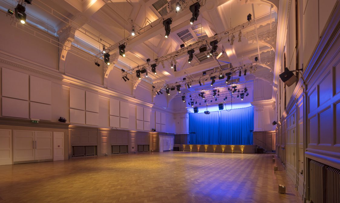 A photograph of the Great Hall, which has a stage with a blue curtain and a lighting rig.