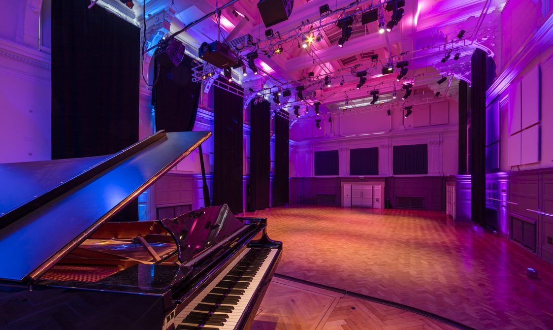A photograph of the Great Hall, with a piano on the floor. There is a lighting rig and purple lighting in the room.