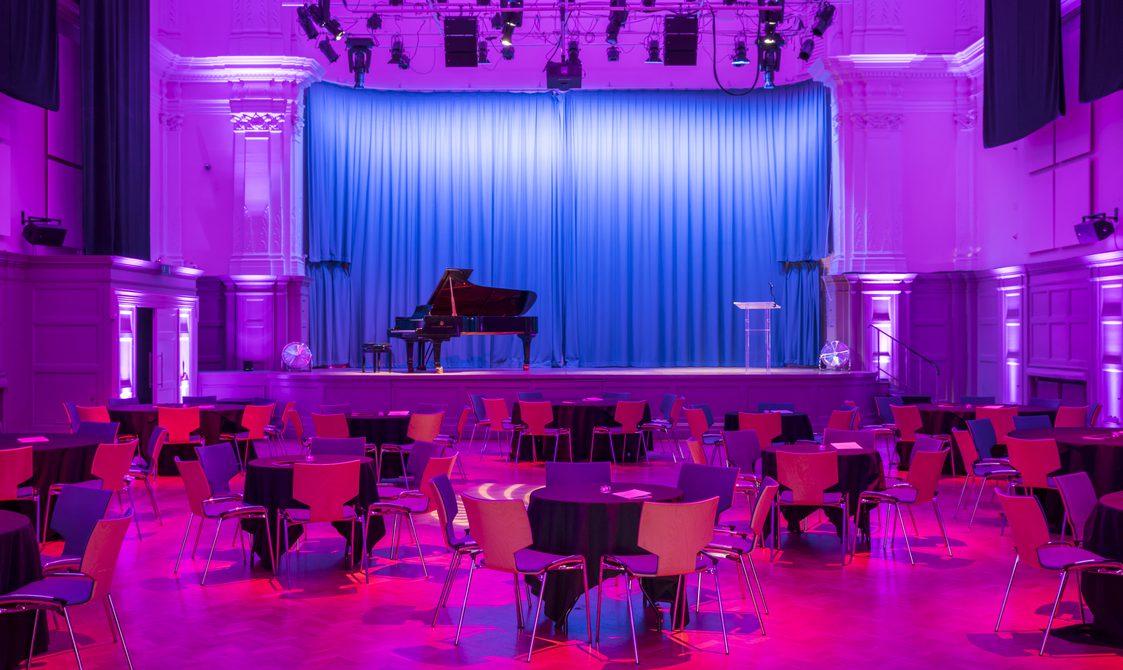 A photograph of the Great Hall, with seating in a cabaret style. There is a stage with a piano on it and blue curtain. There is a purple wash over the room.