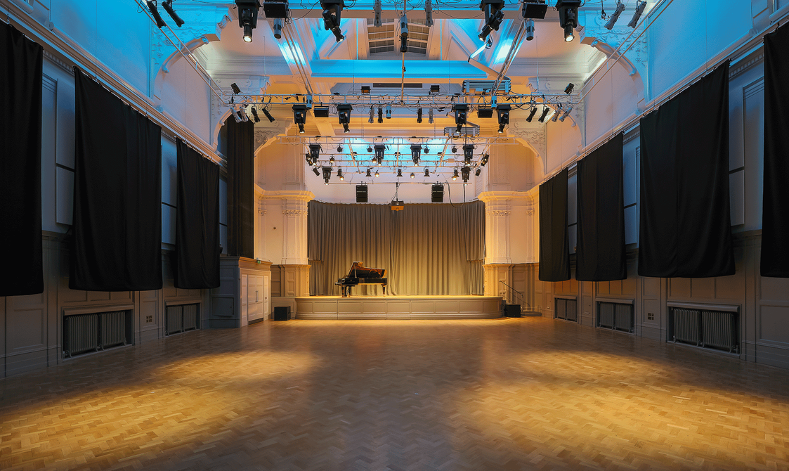 A photograph of Bishopsgate Institute's Great Hall in London. There is a piano on the stage and the lighting rig is producing a yellow wash over the room.