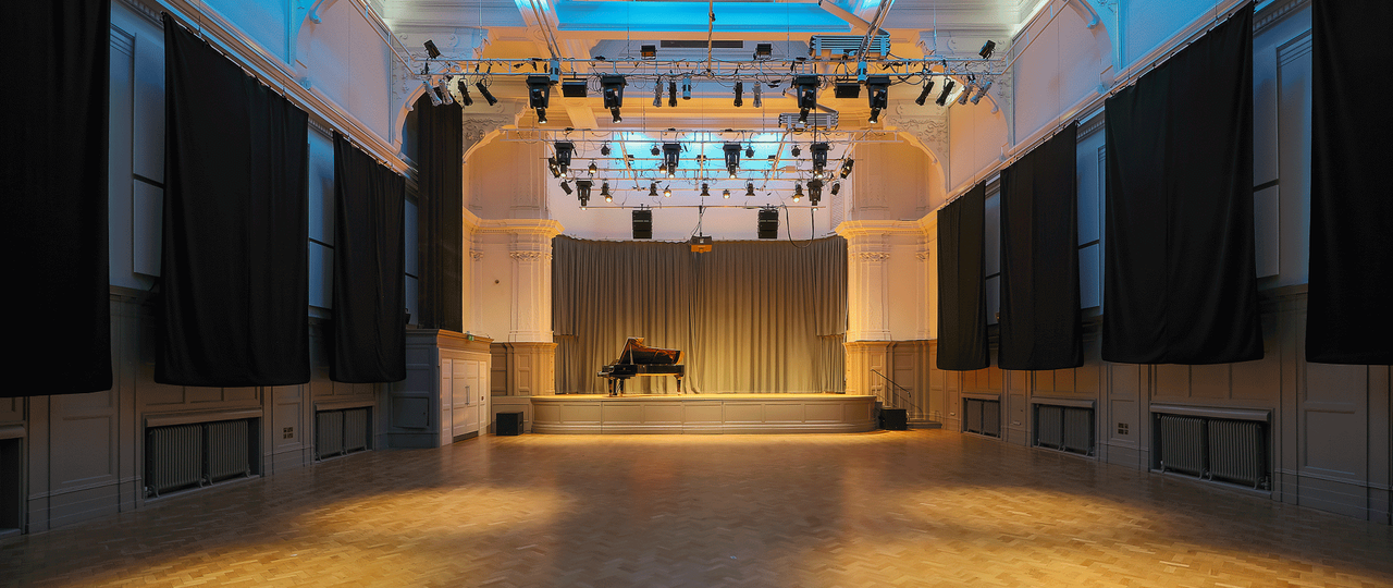 A photograph of Bishopsgate Institute's Great Hall in London. There is a piano on the stage and the lighting rig is producing a yellow wash over the room.