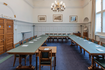 A photograph of the Boardroom at Bishopsgate Institute in London. The tables are set up in a boardroom style with chairs around it. Three landscape pictures are on the wall behind the tables and a chandelier is hanging down.