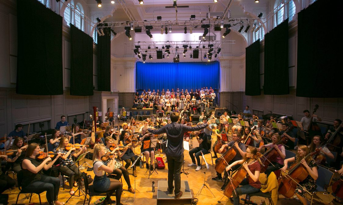 An orchestra rehearsal in Bishopsgate Institute's Great Hall in central London. A conductor is standing in the middle with musicians around them playing the cello, violin, and other instruments. There is a stage at the back of the room.