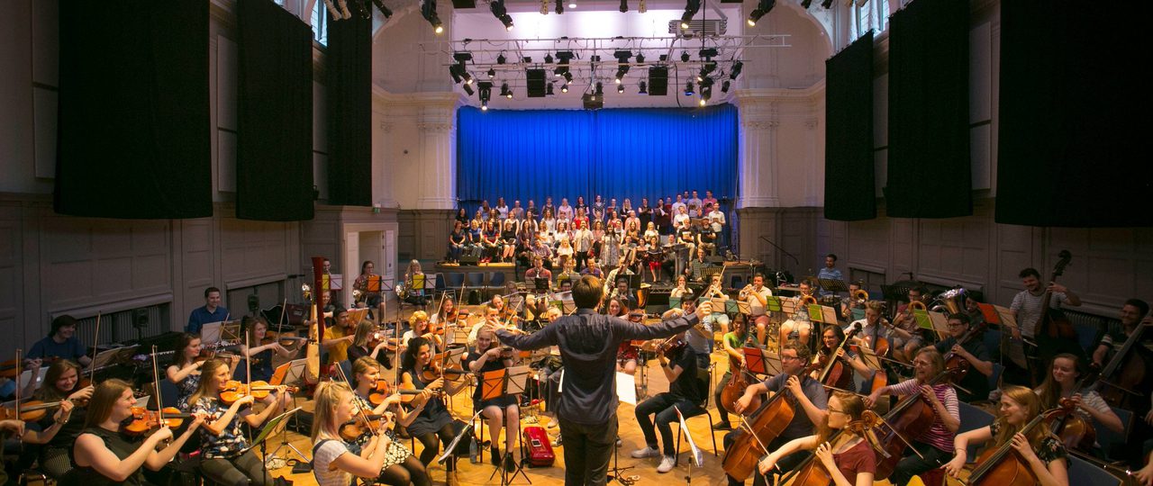An orchestra rehearsal in Bishopsgate Institute's Great Hall in central London. A conductor is standing in the middle with musicians around them playing the cello, violin, and other instruments. There is a stage at the back of the room.