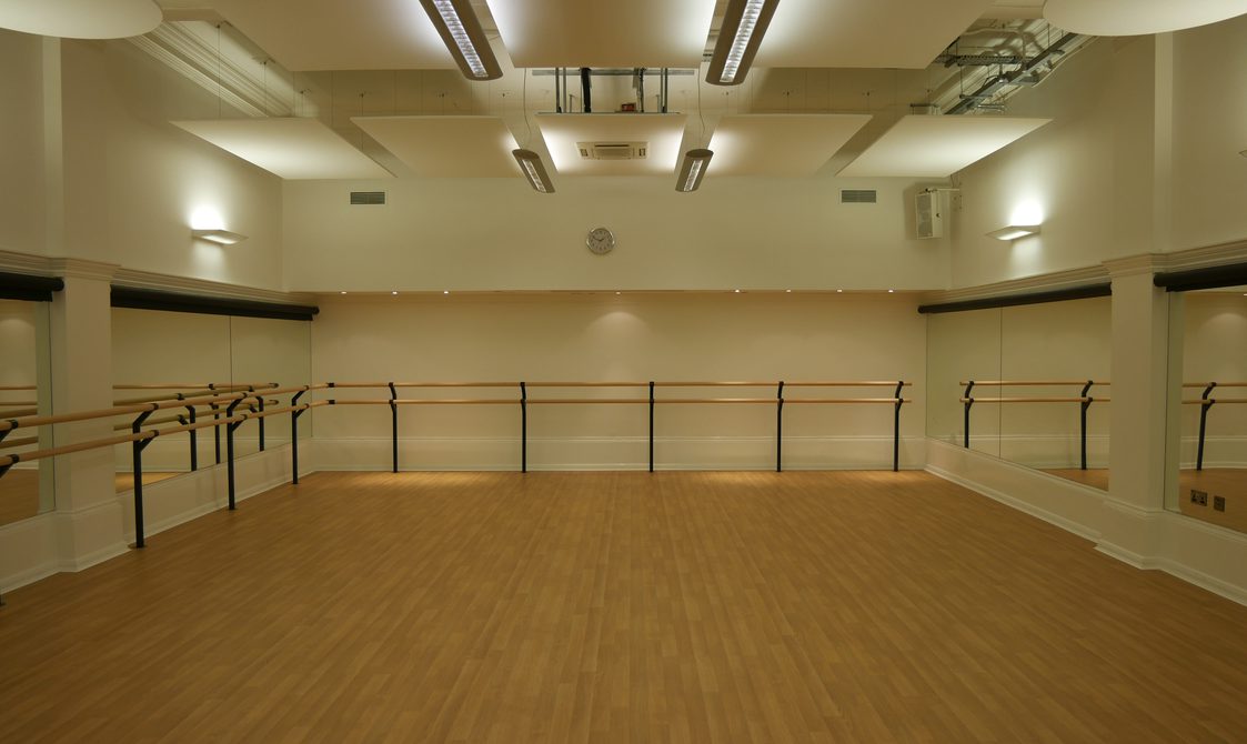 A photograph of the Studio. The floor is wooden and the walls are white, with dance bars around the edge of the room and mirrors on the walls.