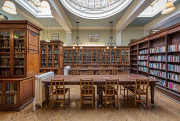 A photograph of a London-based Library. There is a wooden table in the middle of the room with rows of bookshelves around it. There is a dome in the ceiling letting light through.