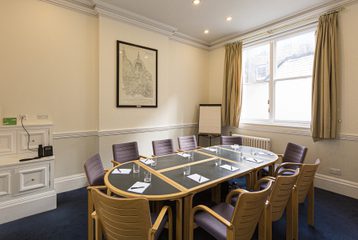 A photograph of the Townsend Room. A conference table is in the middle of the room with 11 chairs around it. There is a window with beige curtains and a print on the wall. The walls are painted white and the carpet is blue.