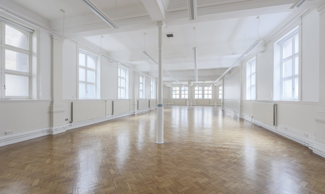 A photograph of the Upper Hall with natural light coming through. There are large windows on the white walls, and the floor is laminated.