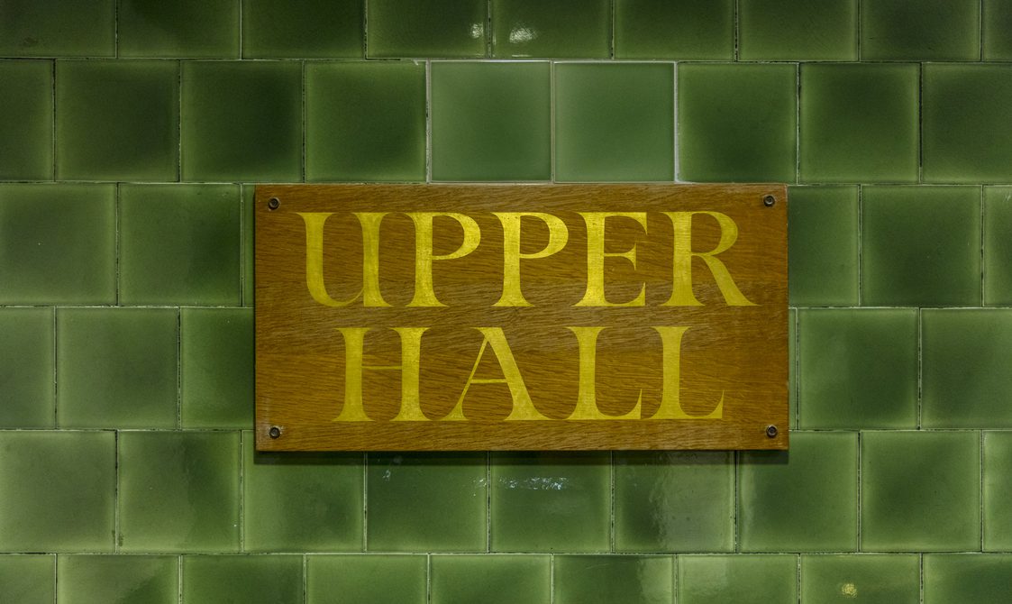 A green tiled wall with a wooden sign with gold writing that says: "Upper Hall"