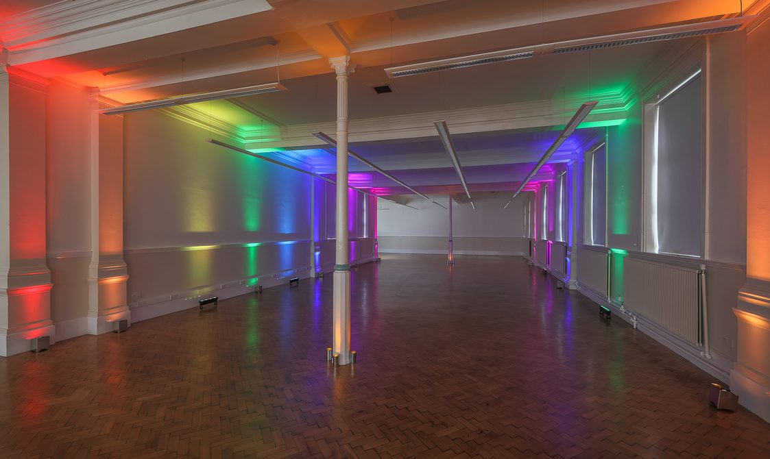 A photograph of the Upper Hall, which has laminate flooring and white walls. Rainbow coloured uplighters are on.