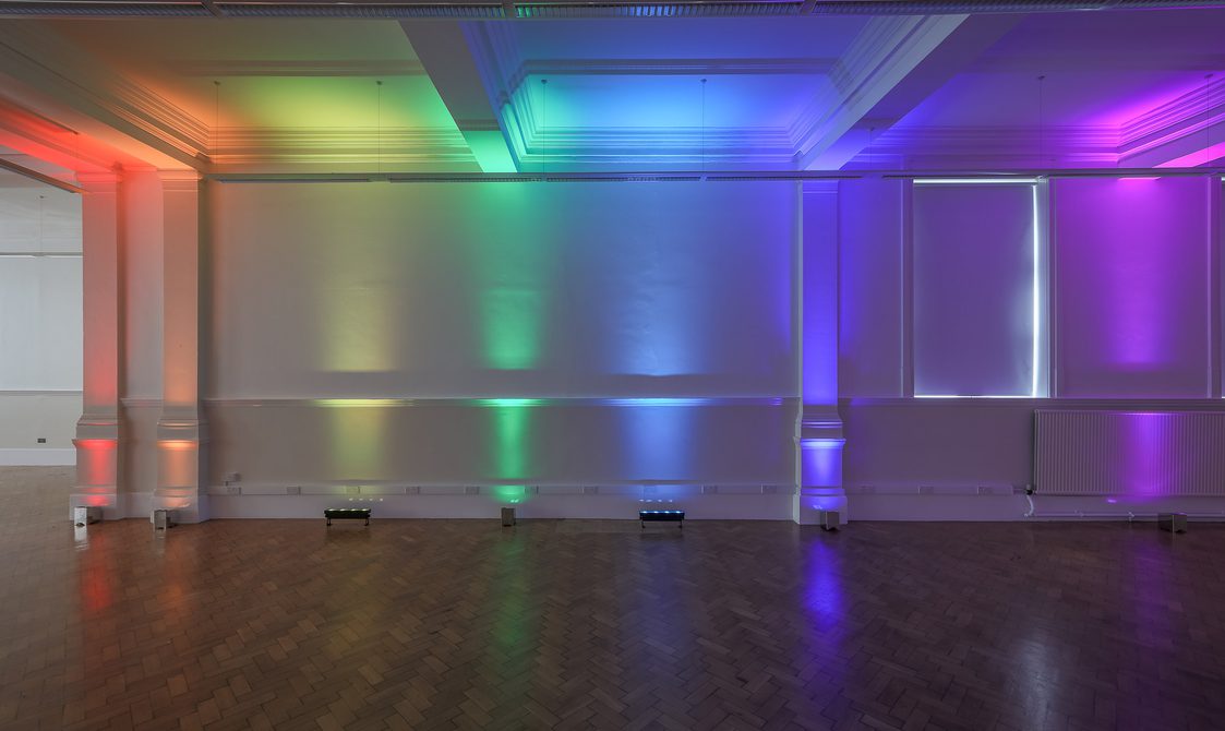 A photograph of the Upper Hall, with has a wooden floor and white walls. The uplighter are on, which are projecting rainbow colours up the wall.