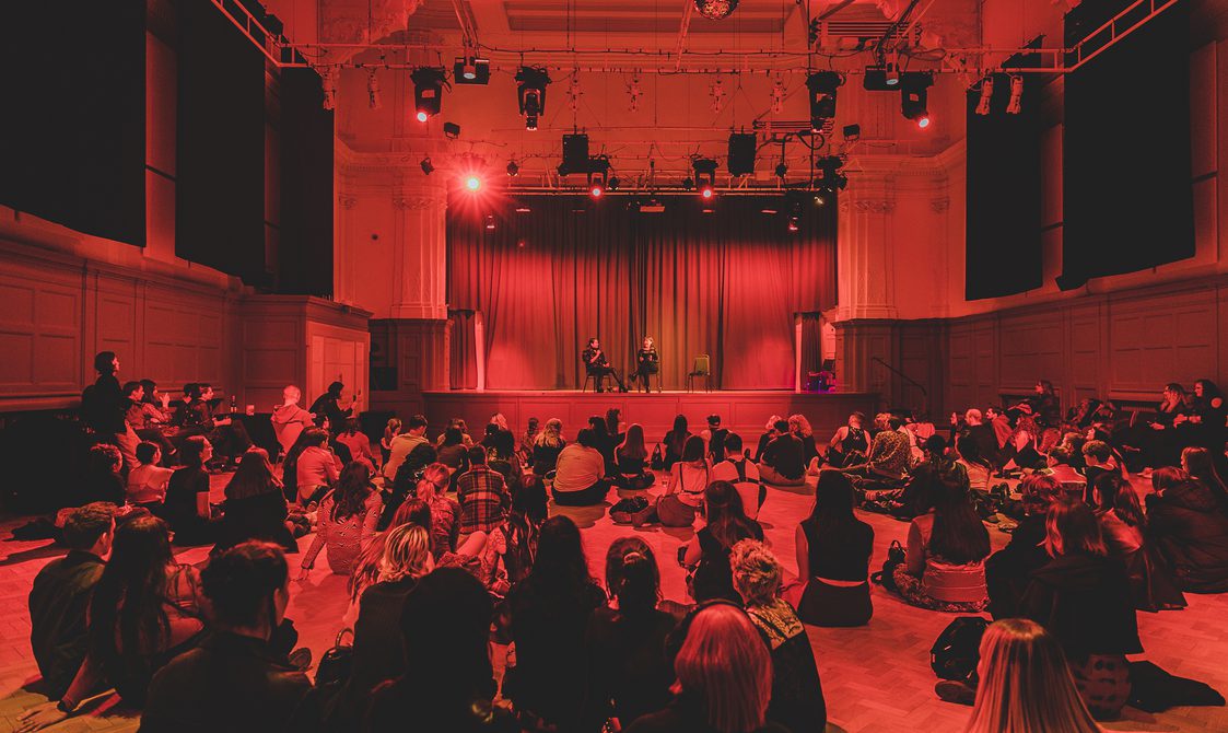 A large group of people sat on the floor of the Great Hall, watching two speakers who are seated on stage