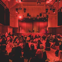 A large group of people sat on the floor of the Great Hall, watching two speakers who are seated on stage