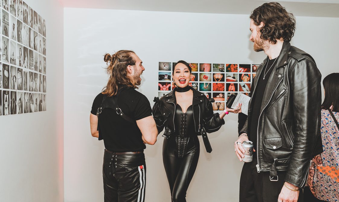 Dominatrix Eva Oh, standing with two other people. All three are dressed in black leather. They are in front of a photography display.