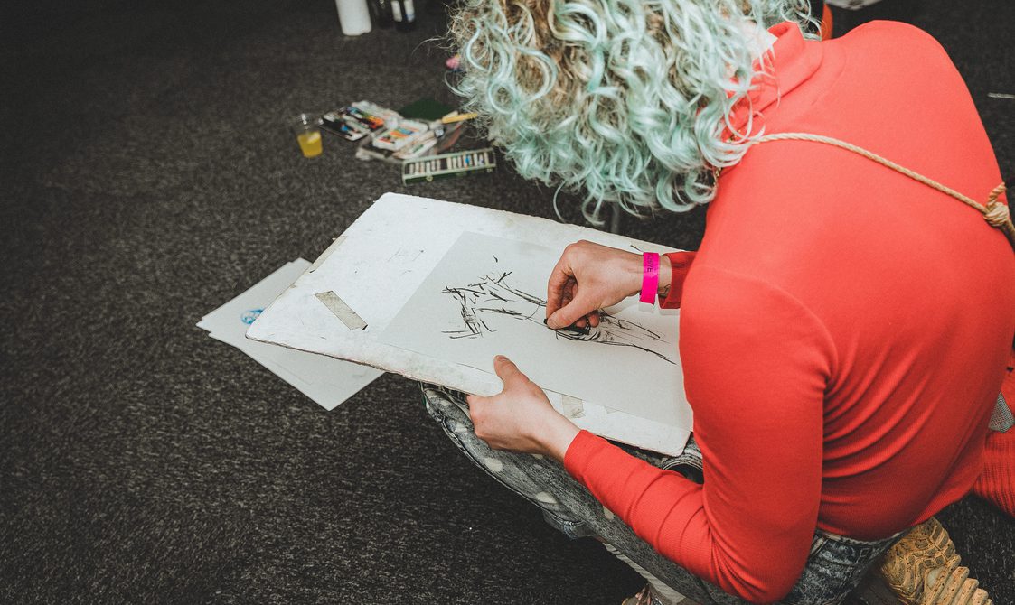 A person with blonde curly hair, wearing a red top, drawing in charcoal