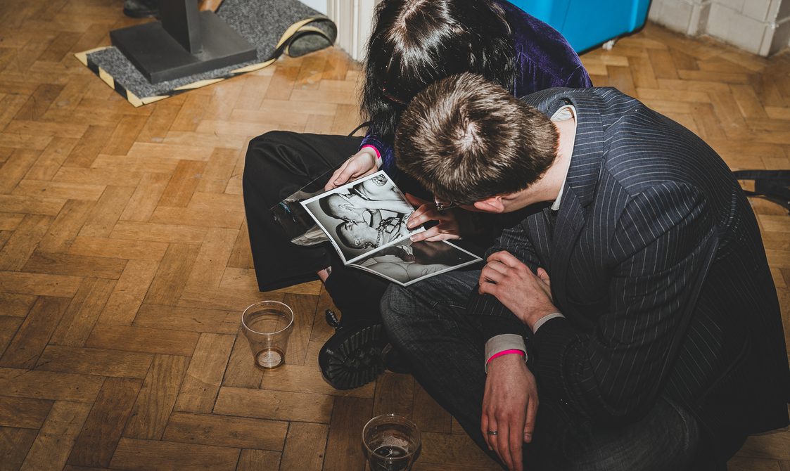 Two people seated on the floor, looking at a book of photographs from the Kink archive. One person wears a pinstripe suit, the other a purple velvet top and black trousers.