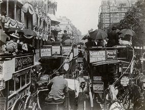 Archive image: A black and photograph of crowded horse-drawn omnibuses travelling down Picadilly. Many passengers on the open-tip buses carry open umbrellas.