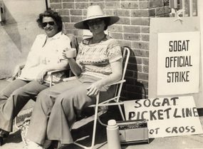 Black and white photograph of two people sitting in deckchairs wearing sunglasses. One has a sunhat and drinks a cup of tea from a thermos. A sign shows they are sat at the SOGAT picket line, as part of the SOGAT Official Strike.