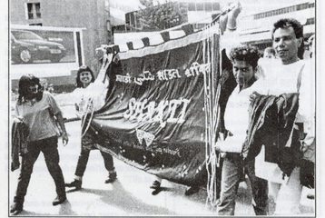 Black and white photo of people marching, holding a large Shakti banner