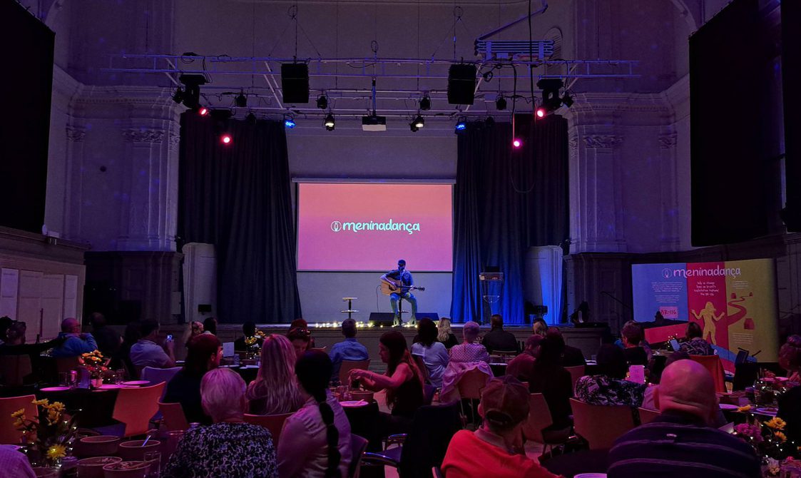 A seated crowd watches a performer, playing guitar and singing, perform on stage in the Great Hall.