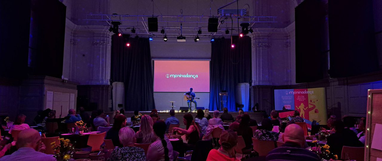 A seated crowd watches a performer, playing guitar and singing, perform on stage in the Great Hall.