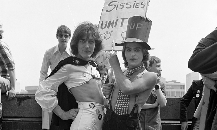 Two people in 1976, wearing non-binary clothes and carrying a sign that says 'Sissies Unite'