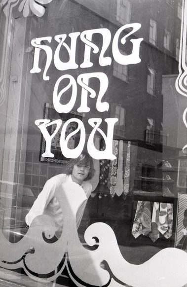 A black and white photo from the 1960s of a man stood inside a shop looking out the window. Stickers on the glass of the window say 'Hung on You'