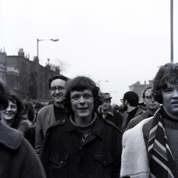 A black and white photo of protesters at the Aldermaston March. A teenage boy is the focus of the picture. Smiling, surrounded by other teens on the march.