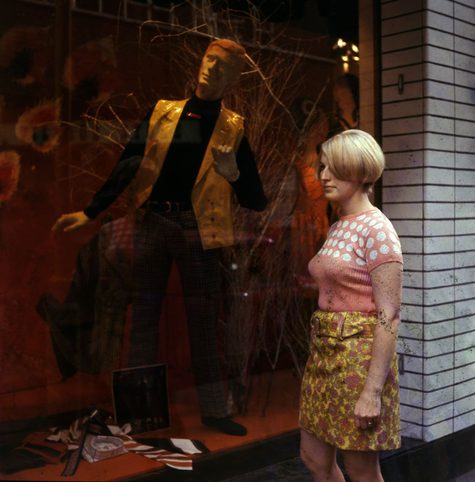 A colour photo from the 1960s of a model stood outside a shop window, imitating the mannequin inside the shop.