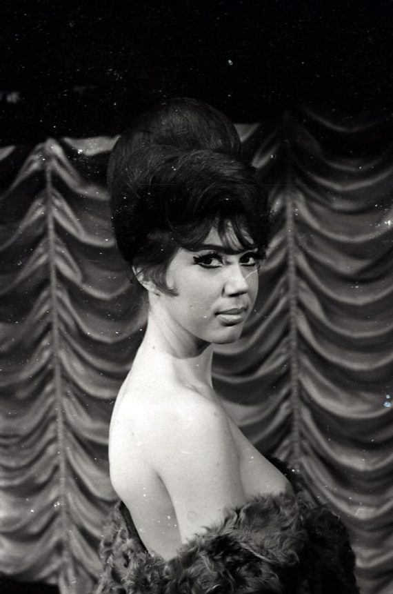 A black and white photograph of a stripper in the 1960s. She is naked, but draped in a fur. She has a large beehive hairstyle and is stood in front of a theatrical curtain.