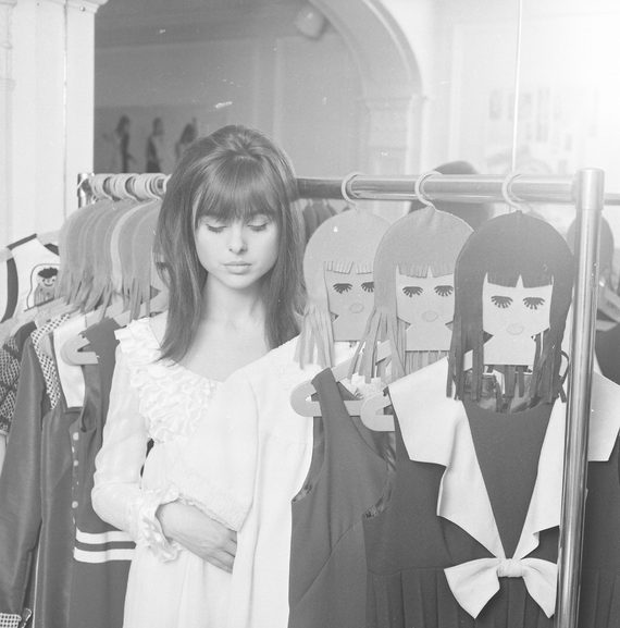 A hazy photo of a model in the 1960s, stood in a shod and imitating the mannequins around her.