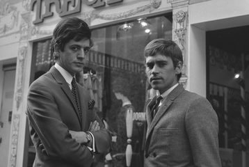 Black and white photo from the 1960s of two young men dressed in suits, with similar mod haircuts, looking stern. They are stood outside a clothing shop called Tre Camp.