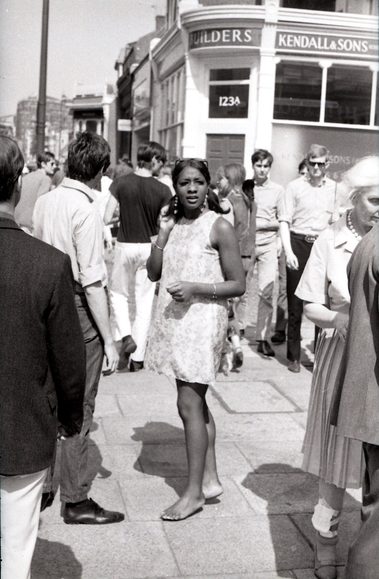 A black and white photo of a model stood on the pavement on King's Road, as people pass her by. She is barefoot and wearing a Mary Quant mini dress