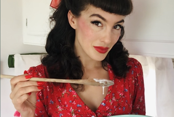 Sugar May holds a mixing bowl and wooden spoon. They are wearing a red dress and have black hair styled in 1940s victory rolls with a red bow